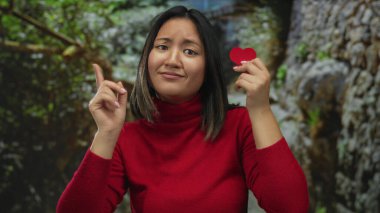 Dışarıdaki genç kadın kırmızı bir kalp tutuyor ve bir parkta dikilirken, duygularını ve düşüncelerini ifade eden kırmızı bir kazak giyiyor..