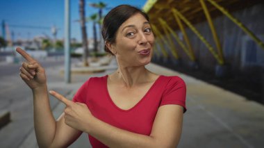 Orta yaşlı, kırmızı tişörtlü, gülümseyen ve güneşli bir şehir sokağındaki palmiye ağaçlarını işaret eden iyimser bir kadın..