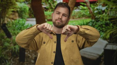 Yeşil bitkiler ve ağaçların arasında, hardal rengi ceket giyen bir adam, park yolunda başparmağını kaldırıyor..