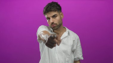 Beyaz tişörtlü genç İspanyol, canlı mor renkli stüdyo ortamında genişletilmiş parmağıyla işaret eder; davet çağrısı.