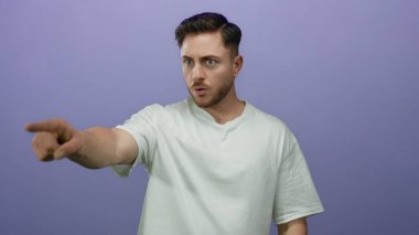 Beyaz tişörtlü genç sakallı adam yoğun bir şekilde izole bir mor duvar arka planı işaret ediyor, odak ve kararlılık ifade ediyor.