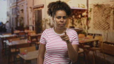 Kadın açık hava restoranının terasında kamerayı işaret ediyor, çizgili gömlek ve yüzük giyiyor, parmağına yüzük takıyor, kıvırcık saçlı, masaların ve sandalyelerin yanında duruyor; kızgın uyarı.