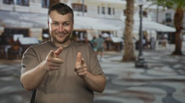 Caddedeki kafede, palmiye ağaçları ve kaldırım desenleri olan bir adam iki parmağını da kameraya doğrultuyor. Arkadaşça bir cazibe..