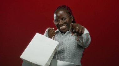 Kırmızı arka planda alışveriş torbalarıyla gülümseyen kadın, neşeli alışveriş deneyimi ve tüketici memnuniyetini yansıtıyor.