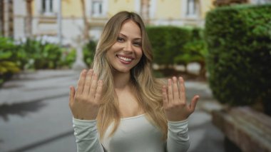 Kadın, kent caddesinde gülümserken iki elini de havaya kaldırır; davet sıcaklığı bağlantısına hoş geldin der.