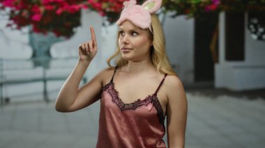 Şehir merkezinde, arka planda renkli çiçekler olan bir sokakta gece elbisesi giyerken maske takan bir kadın görülüyor..