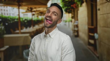 Kentsel sokak ortamında neşeli bir şekilde gülümseyen genç bir adam masaların ve binaların bulanık arka planıyla, gün içinde neşeli ve canlı şehir atmosferini yakalıyor..