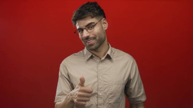 Kırmızı stüdyoda bej gömlekli ve gözlüklü bir adam açık avuç içi jesti ve ince bir gülümseme; merak.