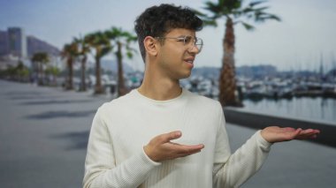 Beyaz süveter giyen ve gözlüklü adam, palmiyelerin palmiye ağaçlarının altında gezindiğini gösteriyor; belirsizlik..