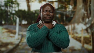 Yeşil kazaklı bir kadın parkta gergin bir ifadeyle boynuna dokunuyor. Bulanık ağaçlar ve patikalarla çevrili nefes almakta güçlük çekiyor..