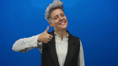 Mavi stüdyoda beyaz gömlek giyen, siyah yelek giyen, gülümseyen ve baş parmak işareti yapan bir kadın..