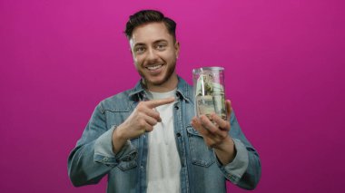 Sakallı genç bir adam, izole edilmiş pembe bir arkaplanı işaret eden bir kavanoz dolusu dolarımızı tutuyor. Kot ceket giyiyor ve kameraya içten bir şekilde gülümsüyor.