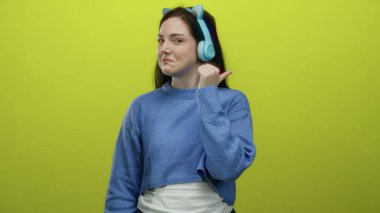 Mavi süveter ve kulaklıkla gülümseyen kadın, parlak sarı arka plana başparmağıyla işaret ediyor, neşeli bir ifade ve modern bir tarz sergiliyor..