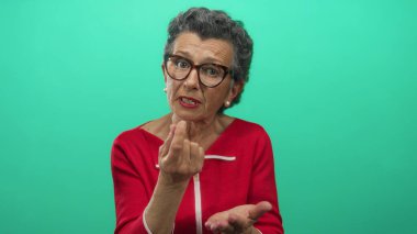 Kırmızı tişörtlü, gri saçlı yaşlı bir kadın yeşil arka planda para için el kol hareketi yapıyor. Zorlayıcı bir görsel tasvirle maddi gereksinim ve bağımsızlık taşıyor..