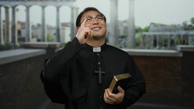 Gözlüklü ve dini kıyafetleri olan Asyalı rahip İncil 'i tutuyor. Arka planda tarihi mimarisi olan bir şehir ortamında düşünceli bir şekilde el kol hareketi yapıyor..