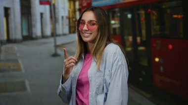 Pembe tişörtlü ve güneş gözlüklü genç bir kadın, şehir sokaklarında gülümsüyor. Kırmızı otobüslerin ve binaların arka planında canlı şehir atmosferini gösteriyor..
