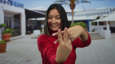 Kırmızı kazaklı kadın gülümsüyor. Canlı şehir caddesinde palmiye ağaçlarıyla hashtag jesti yapıyor. Açık havada neşeli şehir hayatı sergiliyor..