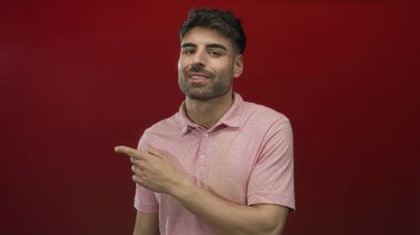 Kırmızı stüdyoyu işaret eden pembe polo tişörtlü ve gülümseyen İspanyol genç adam güven güçlendirme pozitifliği.