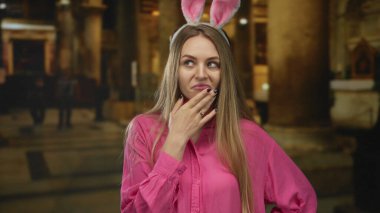 Pembe gömlekli ve tavşan kulaklı bir kadın kilise ortamında neşeli bir ifade sergiliyor. Dini arka planda tuhaf kıyafetlerini vurguluyor..