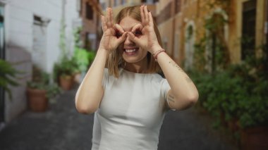 Elleriyle dürbün hareketi yapan genç bir kadın, canlı bir şehir ortamında kırsal binaları ve yeşillikleri olan eski bir kasaba caddesinde gülümsüyor..