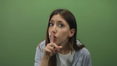 Çizgili gömlek giyen genç İspanyol kadın ve küpeler stüdyo ortamında sessizlik için parmakları dudaklara işaret ediyor; sessizlik sessizlik.