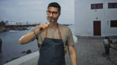 Önlüklü ve gözlüklü bir adam, sahil kenarındaki bir binanın yanında iki kez baş parmak hareketi yapıyor. Onaylanmıyor..