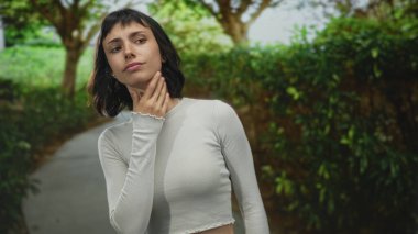 Kısa siyah saçlı, beyaz uzun kollu, yeşil ormanda boynuna dokunup gözlerini kapatan bir kadın..