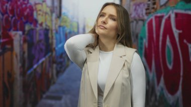 Şehir caddesinde bej ceketli, boynu rahatsız bir şekilde boynu tutarak renkli grafiti yapan bir kadın, şehir yaşam tarzını öneriyor..