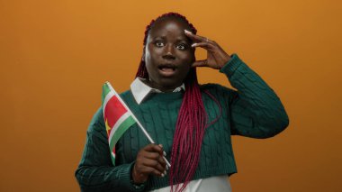 Turuncu arka planda surinam bayrağı olan bir kadın yeşil kazak ve pembe örgüler giyerken düşünceli bir jest yapıyor..