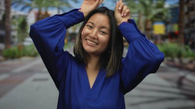 Woman making playful horns gesture in blue blouse while smiling outdoors on a city street with palm trees in the background capturing a cheerful urban vibe.