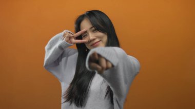 Confident young woman in sweater pointing forward against isolated orange wall posing playfully with a cheerful expression.
