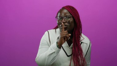 Gözlük ve beyaz süveter giyen, soyutlanmış pembe bir arka plana karşı sessiz kalmak için el kol hareketi yapan Afrikalı Amerikalı bir kadın..