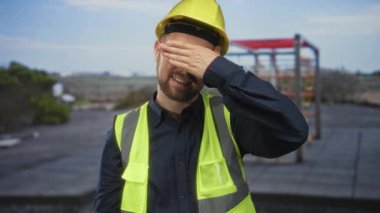 Sarı kasketli ve çelik yelek giyen insan müteahhiti, bina iskelesinin yakınındaki çatıda eli ile gözlerini kapatıyor..