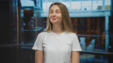 Genç bir kadın otel ortamında gülümsüyor, beyaz bir gömlek giyiyor, bir yandan bakarken mutluluk ve sükunet anı yakalıyor..