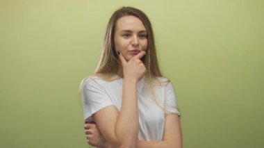 Beyaz gömlekli genç bir kadın, izole edilmiş sarı arka planda, düşünme yüzüyle, çene üzerinde elle kafa yorarak, merak ve içgözlem yapıyor..