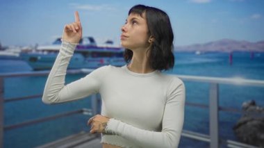 Beyaz mahsullü kadın gün ışığında deniz yatı ile güneşli iskelede yukarı doğru işaret ediyor; gelecekteki iyimserlik.