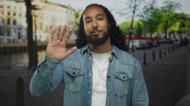 Kameraya bakarken beyaz tişört ve kot ceket giyen bir adam sokak kanalında el sallıyor. Dostluk selamlaşması..