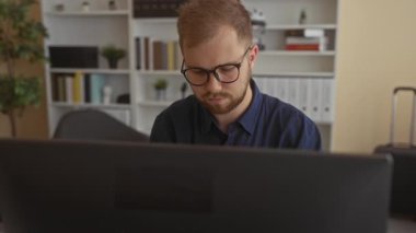 Gözlüklü genç beyaz adam ofis binasında otururken işaret parmağıyla bilgisayar monitörüne işaret veriyor..