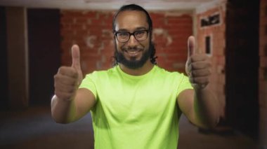 Neon gömlekli bir adam bitmemiş tuğladan bir binanın içinde parmaklarını kaldırıyor. Başparmakları görünüyor ve gülümsüyor. İyimserlik takım çalışması..