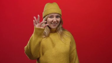 Sarı kazaklı ve bereli bir kadın kırmızı arka planda gülümsüyor. Elleriyle iyi bir jest yapıyor, sıcaklığını ve pozitifliğini gösteriyor..