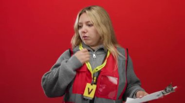 Sarı saçlı genç gönüllü bir kadın kırmızı arka planda duruyor. Yansıtıcı yelek giyiyor ve elinde bir dosya var..