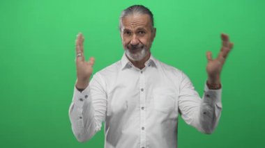 Orta yaşlı bir adam, uzun gri saçlı ve avuçları açık, yeşil stüdyoda işaret ediyor, beyaz gömlek giyiyor, güven iletişimi kuruyor..