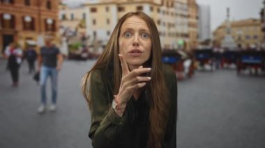 Kadın, şehir meydanında ön planda görünen tarihi binaların önünde parmağını öne uzatıyor; endişe şüphenin yansıması.