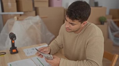 Adam faturaları biriktirip evdeki toplamları sayarken genç bir İspanyol masanın yanındaki yeni kutuların yanında para izi sürüyor..