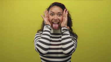 Genç İspanyol kadın elleri ile yüzünü örtüyor, avuçlar gözlerin üzerinde ve çizgili üst sarı bir stüdyo ortamında; oyuncu neşe.