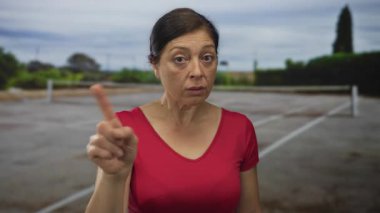 Kırmızı tişörtlü orta yaşlı bir kadın güneş ışığı altında açık tenis kortunda işaret parmağıyla kameraya işaret ediyor..