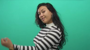 Genç İspanyol kadın, yeşil stüdyoda siyah beyaz çizgili bluz giyerek kol kola, alnına kadar esnedi; kendine güveni güçlü neşe.