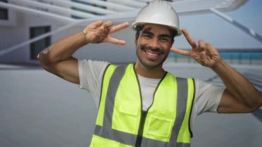 Kasketli ve yansıtıcı yelekli bir adam modern bina girişinde iki eliyle barış işareti yapıyor..