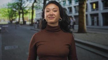 Yüzü açık gülümseyen, parmağıyla işaret eden ve halka küpeli bir kadın, caddede kahverengi balıkçı yaka giyiyor; neşeli bir bağlantı..