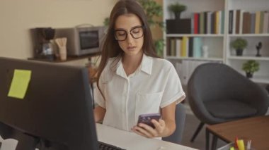 Ofis binasında akıllı telefon tutan ve klavyeye yazan, gözlük takan ve bilgisayar monitörüyle masaya oturan kadın, konsantrasyon verimliliği.